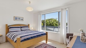 A bedroom with a wooden bed, a large window with a view of greenery, and a small table with a vase. - Dolphin Dreaming - 16 Dolphin Avenue (CARRICKALINGA)