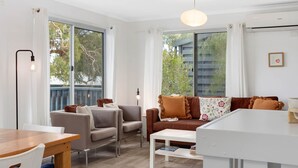 Ein modernes Wohnzimmer mit einer Couch, Sesseln, einem Esstisch und großen Fenstern mit Blick auf Bäume und Gebäude.