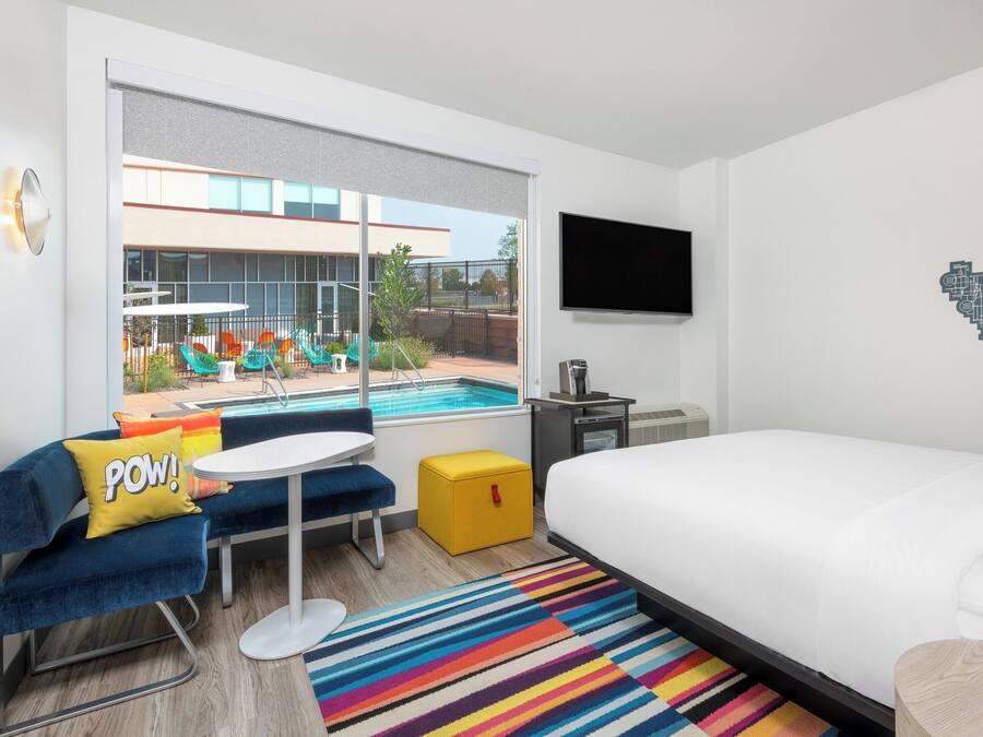 A modern hotel room with a large window, a bed, a small table, a chair, a TV, and a pool view.