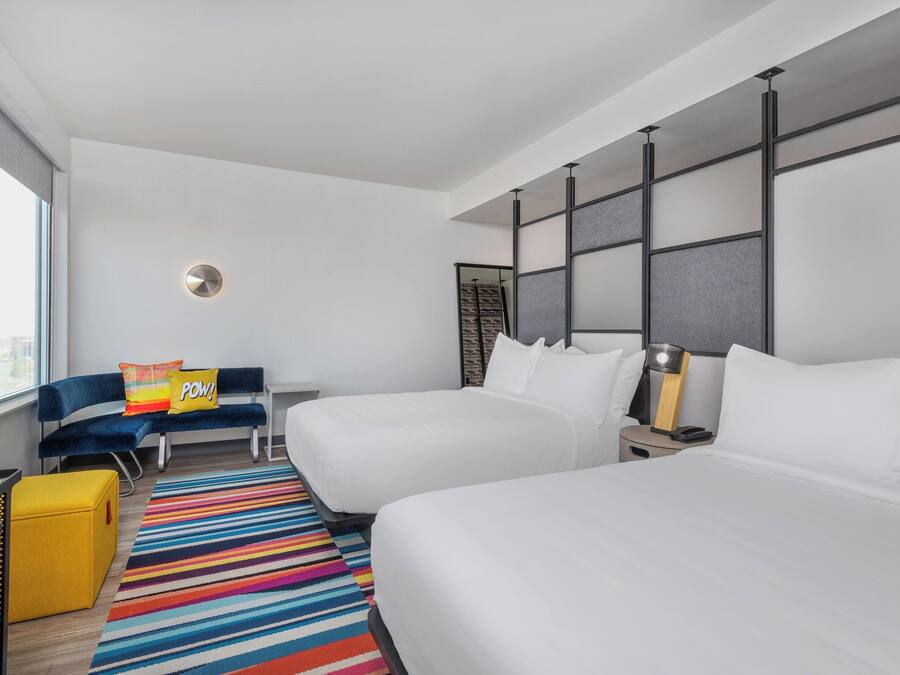 Une chambre d’hôtel moderne avec deux lits, un canapé, un tapis rayé aux couleurs vives, un petit pouf jaune et une grande fenêtre donnant sur la ville.