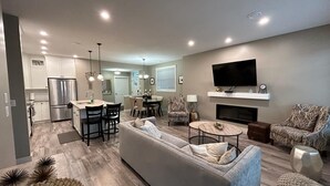 A modern living room with a grey sofa, a TV mounted on the wall, and a kitchen in the background. - OSOYOOS PRIVATE RESORT HOME STEPS FROM THE LAKE (Oliver)