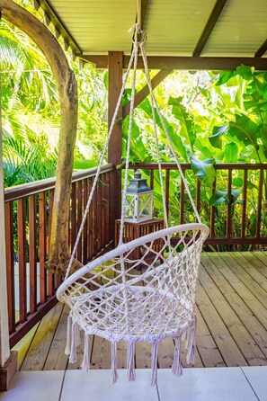 A white hanging chair on a wooden deck with a railing, surrounded by lush greenery.