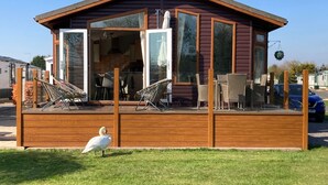 A wooden cabin with a deck, large windows, and outdoor furniture. - River Retreat Lodge is front line with panoramic views over the river Avon. (Stratford-upon-Avon)