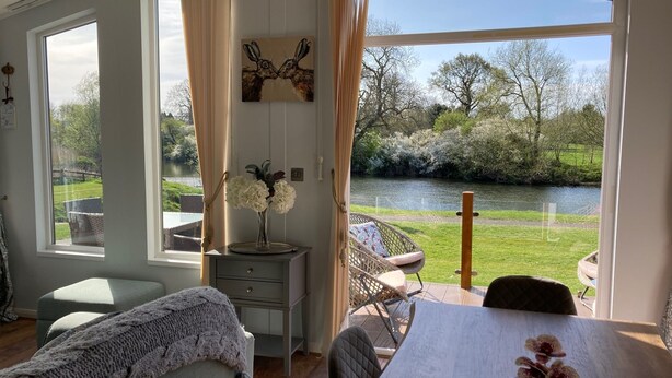 A room with a large window overlooking a river, a dining table with chairs, a sofa, a small cabinet, and a decorative painting. - River Retreat Lodge is front line with panoramic views over the river Avon. (Stratford-upon-Avon)
