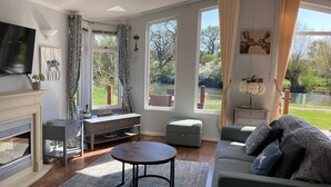 A modern living room with a fireplace, a television, a sofa, a coffee table, and large windows offering views of a garden and a lake. - River Retreat Lodge is front line with panoramic views over the river Avon. (Stratford-upon-Avon)