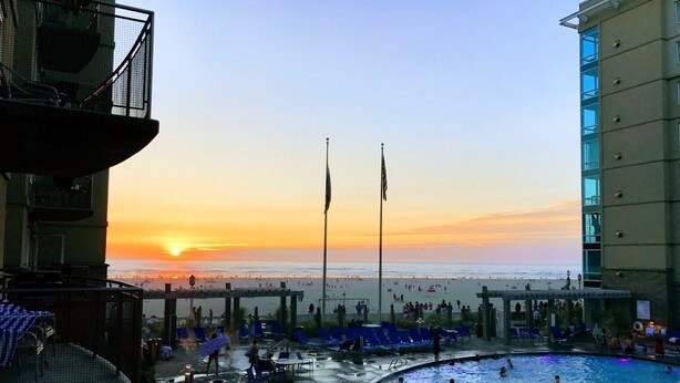 Un espace piscine avec vue sur le coucher de soleil, deux drapeaux et une plage en arrière-plan.