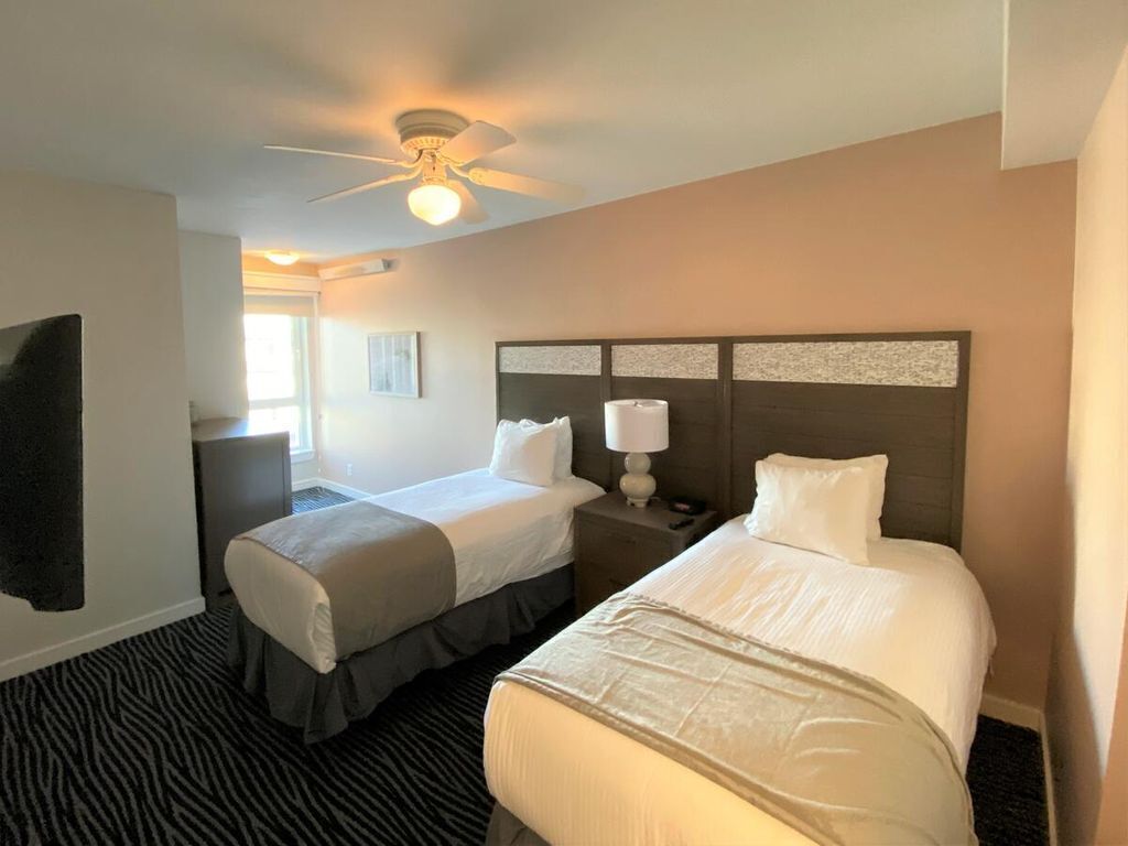 Ein Hotelzimmer mit zwei Betten, einem Deckenventilator, einem Nachttisch mit Lampe und einem Fenster mit Jalousien.