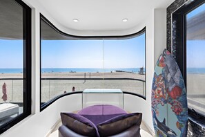 Une chambre moderne dotée d’une grande fenêtre donnant sur une plage, d’une planche de surf et d’une petite table avec un coussin violet.