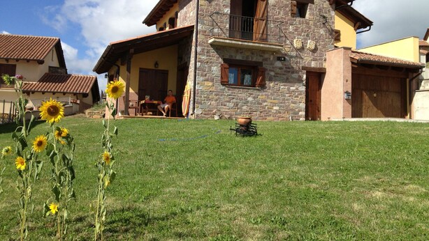 A stone house with a balcony, a grassy lawn, and a sunflower in the foreground.