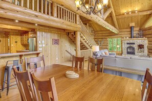 Interior de una cabaña de madera con una mesa de comedor, sillas, una escalera, chimenea y una sala de estar con sofá.