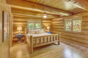 Un dormitorio tipo cabaña de troncos con una cama de madera, dos mesitas de noche, un ventilador de techo y ventanas con vistas al follaje.