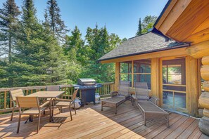 Una terraza de madera con mobiliario exterior, una parrilla y vistas a un bosque.