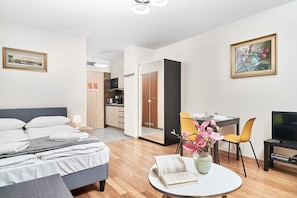 A modern hotel room with a bed, bedside table, a round table with a book and flowers, a TV, and a dining area with chairs.