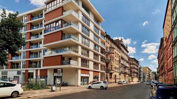 Un immeuble à plusieurs étages doté de balcons, un restaurant bien visible appelé « GROSS », et une rue bordée de voitures garées.