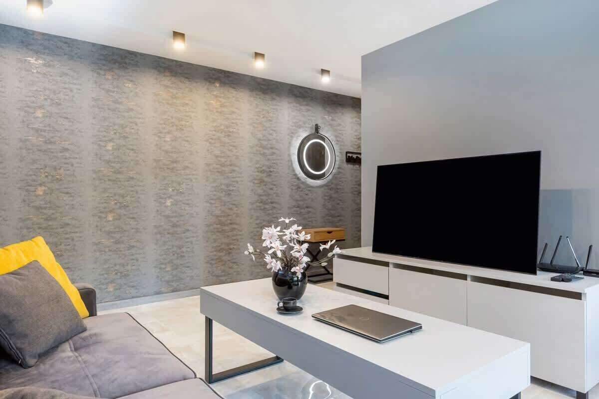 Uma sala de estar moderna com um sofá cinzento, uma mesa de centro branca com um vaso e flores, uma TV de tela plana e um design minimalista.