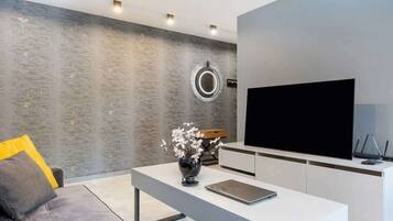 Uma sala de estar moderna com um sofá cinzento, uma mesa de centro branca com um vaso e flores, uma TV de tela plana e um design minimalista.