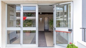 Entrada de un edificio moderno con puertas de vidrio, un pequeño espacio exterior con plantas y un ascensor a la vista.