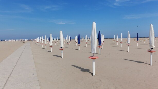 Una playa con hileras de sombrillas blancas y bases rojas, un paseo marítimo de concreto y un cielo azul despejado.