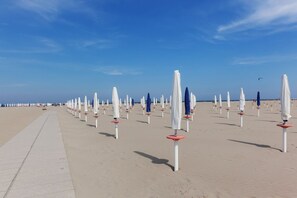 Una playa con hileras de sombrillas blancas y bases rojas, un paseo marítimo de concreto y un cielo azul despejado.