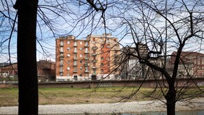 A multi-story building with balconies, a river in the foreground, and trees framing the view.