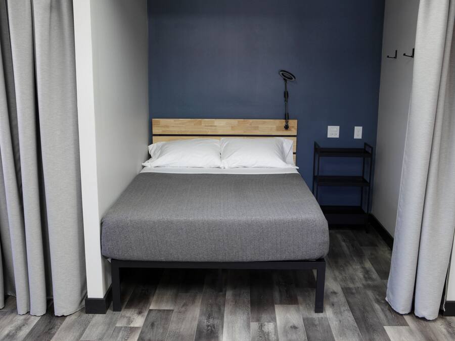 Cama bem arrumada com colchão cinzento e almofadas brancas, cabeceira de madeira, mesa de cabeceira pequena com abajur e cortinas cinzentas.