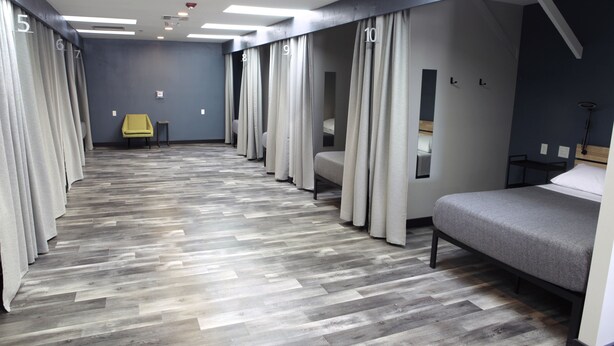 A hotel room with a herringbone-patterned floor, a bed, a chair, and curtains with room numbers. - Stay Classy Hostel (San Diego)