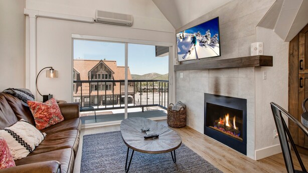 A living room with a fireplace, a sofa, a coffee table, and a large window overlooking a building. - Steps to Shopping, Restaurants, Year-Round Recreation & More! Park City Resort Village 208 (Park City)