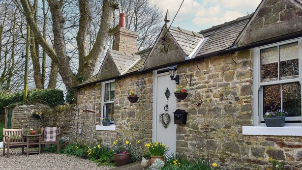 Une petite maison en pierre avec une porte blanche, un petit porche doté d’un banc et des pots de fleurs placés sur le sol.