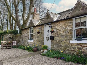 A stone cottage with a white door, a small porch with a bench, and flower pots on the ground. - 1 bedroom accommodation in Thorngrafton, near Bardon Mill (Thorngrafton, near Bardon Mill)