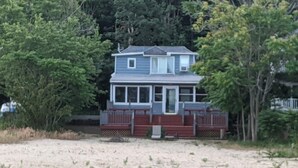 A two-story house with a front porch and a deck, surrounded by trees and a sandy area. - Beachfront Summer Rental- Sea Cliff (Sea Cliff)