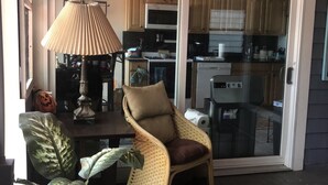 A cozy living area with a wicker chair, a table with a lamp, and a large plant. A kitchen is visible through an open door. - Beachfront Summer Rental- Sea Cliff (Sea Cliff)