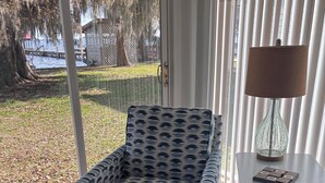 A room with a patterned armchair, a lamp, and a view of a dock and trees through a glass door. - Lakeside Retreat With Panoramic Views Like No Other (Lake Waccamaw)