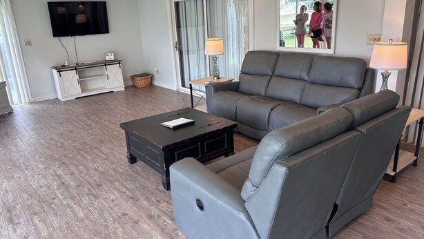 A living room with a grey sofa, a coffee table, a TV, and a window with a view of people outside. - Lakeside Retreat With Panoramic Views Like No Other (Lake Waccamaw)