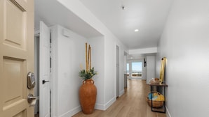 A hallway with a wooden floor, white walls, a large vase with plants, and a small table with a basket. - Direct Oceanfront 2 Bedroom 2 Bathroom on the 1st Floor (Imperial Beach)