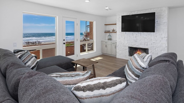 A modern living room with a large gray sofa, a wooden coffee table, and a fireplace. There are large windows offering a view of the ocean. - Direct Oceanfront 2 Bedroom 2 Bathroom on the 1st Floor (Imperial Beach)
