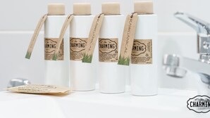 Cuatro botellas blancas con etiquetas y colgadores marrones, identificadas como "The Charming Concept", sobre una superficie blanca en un baño con un grifo moderno.