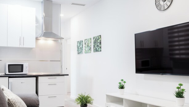 Cocina moderna con gabinetes blancos, microondas y horno integrado. Un sofá blanco con una mesita de centro y dos plantas en macetas. Un televisor de pantalla plana está montado en la pared sobre una consola blanca.