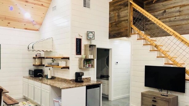 Un espacio habitable compacto con área de cocina, una pequeña mesa de comedor, un televisor integrado y una escalera de madera.