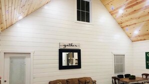 Un acogedor espacio tipo loft con techo inclinado, un sofá de cuero marrón, una mesa de comedor con sillas y un cartel que dice 'gather' sobre un espejo.