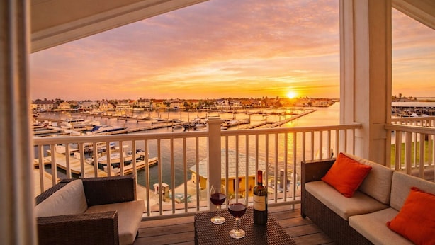 En balkon med fletmøbler, et bord med vinglas og en flaske, samt udsigt til en lystbådehavn i solnedgang.