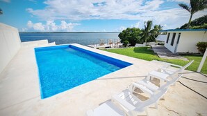 Una piscina rectangular con agua azul cristalina, rodeada de sillones blancos y una zona ajardinada con vista al océano.