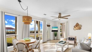 A spacious living room with a large window, a ceiling fan, and a television. - Destiny Beach Villas 2B - Gulf Views, Community Pool, Steps to the Beach! (Destin)