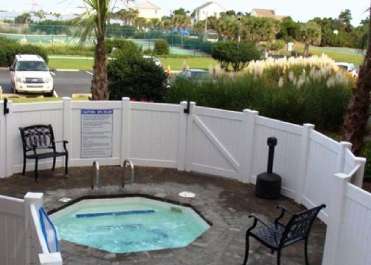 Un espace extérieur aménagé avec un jacuzzi, une clôture blanche, deux chaises en métal noir et un banc.