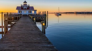 A wooden pier leading to a white building with a red roof, likely a lighthouse, at sunset. - Downtown Manteo with Secret Garden (Manteo)