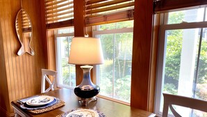 A dining table with placemats, a table lamp, and a view of greenery through large windows. - Downtown Manteo with Secret Garden (Manteo)