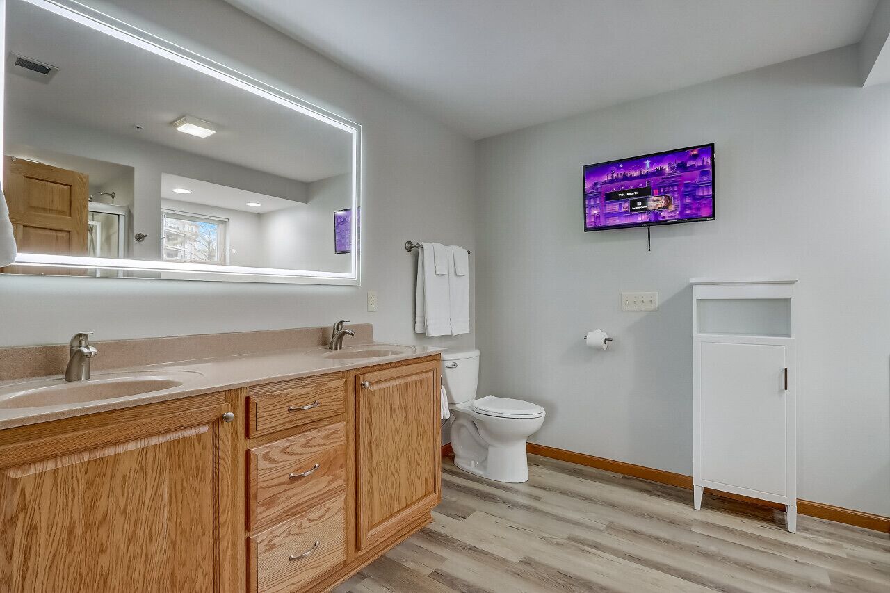 A modern bathroom with a vanity, sink, mirror, toilet, and a television mounted on the wall.