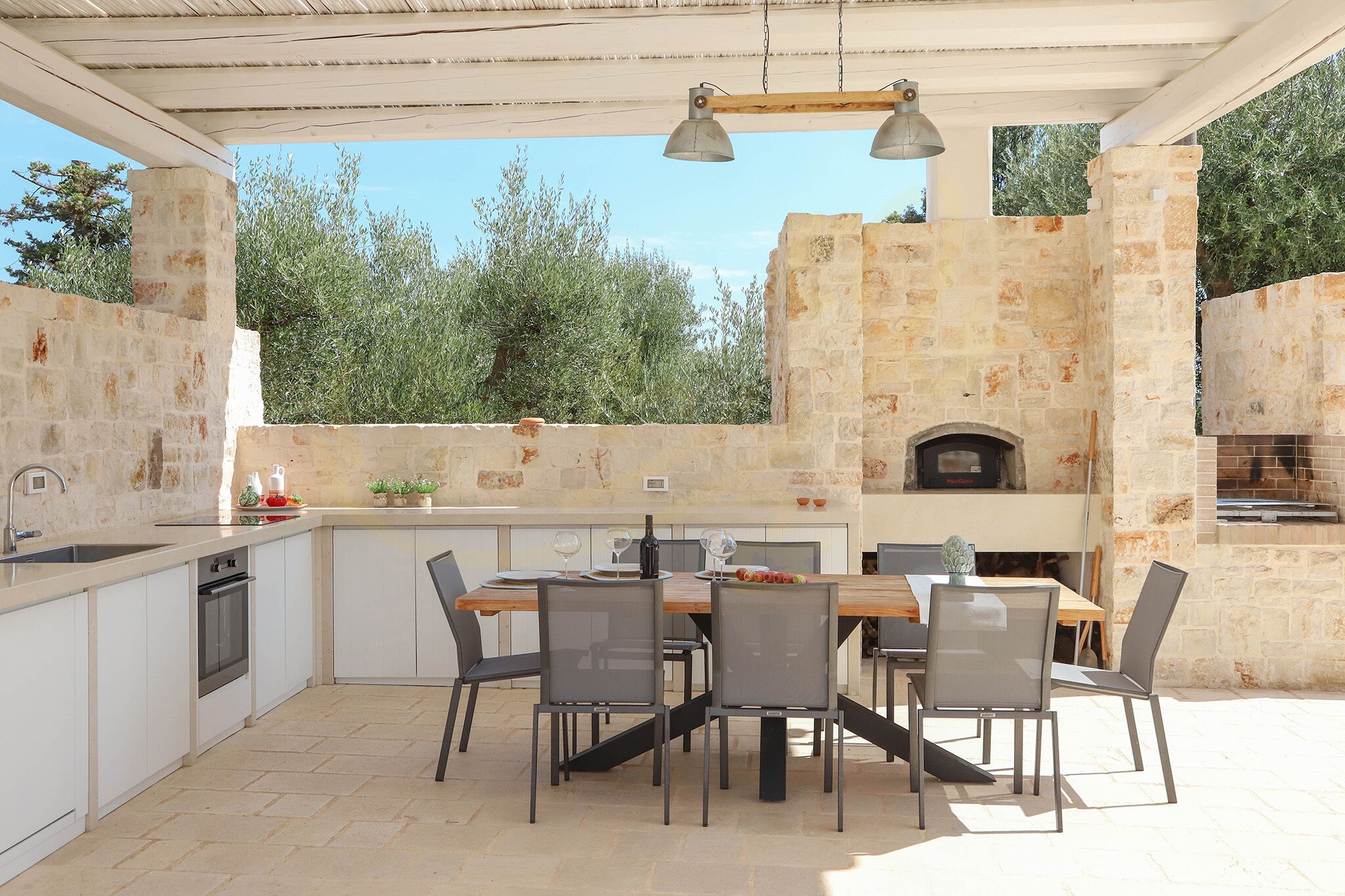 An outdoor kitchen with a stone wall, a built-in oven, and a dining area with chairs and a table.