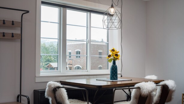 A dining area with a wooden table, two chairs with white fur trim, a vase with sunflowers, and a geometric pendant light.