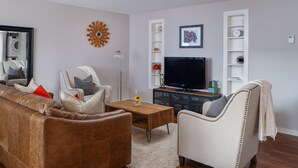 A living room with a brown leather sofa, a white armchair, a wooden coffee table, and a flat-screen TV on a stand.
