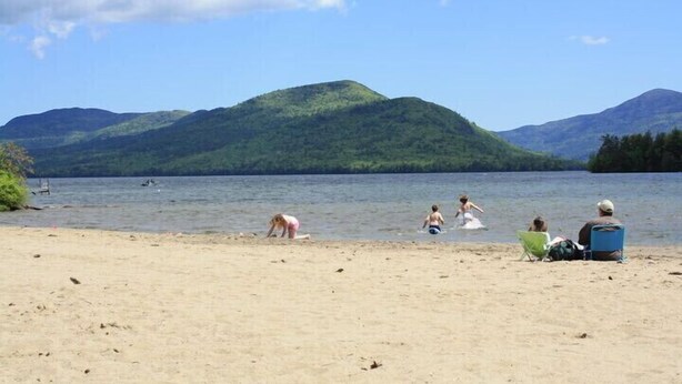 A beach with people engaging in various activities, a clear sky, and mountains in the background. - Lake view cozy two bedroom in Bolton Landing -  walk to Town! (Bolton)